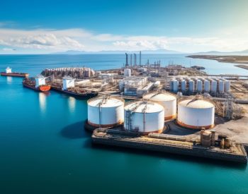 Aerial view of an industrial port with large cylindrical storage tanks, tanker ships, and a sprawling complex by the ocean under a clear blue sky.