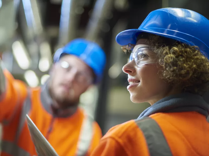 two industrial service engineers conduct a safety check of a control panel and boiler room at a power station . They are both wearing safety equipment and are looking at a control panel .